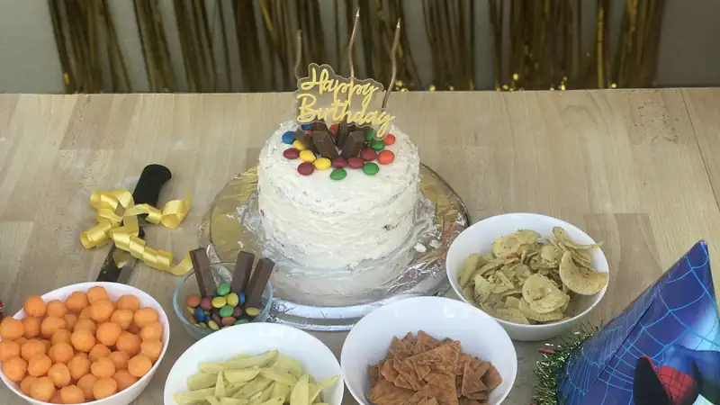 A homemade birthday cake surrounded by bowls of snacks and a party hat on a table.