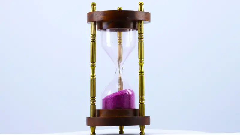 A brass and wood hourglass with pink sand on a plain white background.