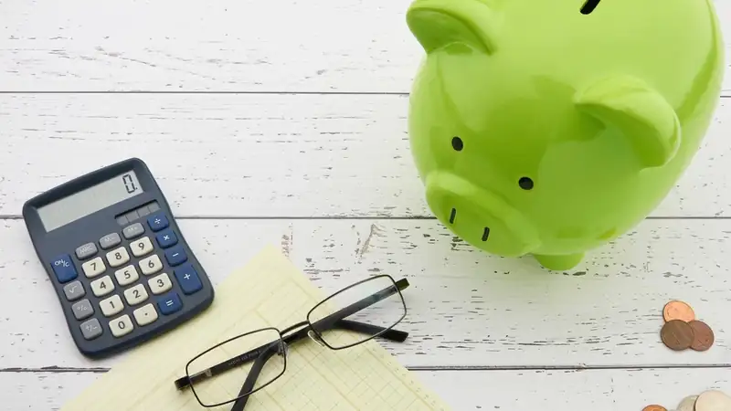 A green piggy bank beside a calculator, reading glasses, a ledger pad and coins on a white table.