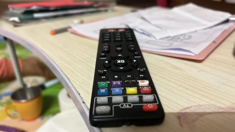 A black TV remote on a wooden table with papers and a mug blurred behind it.